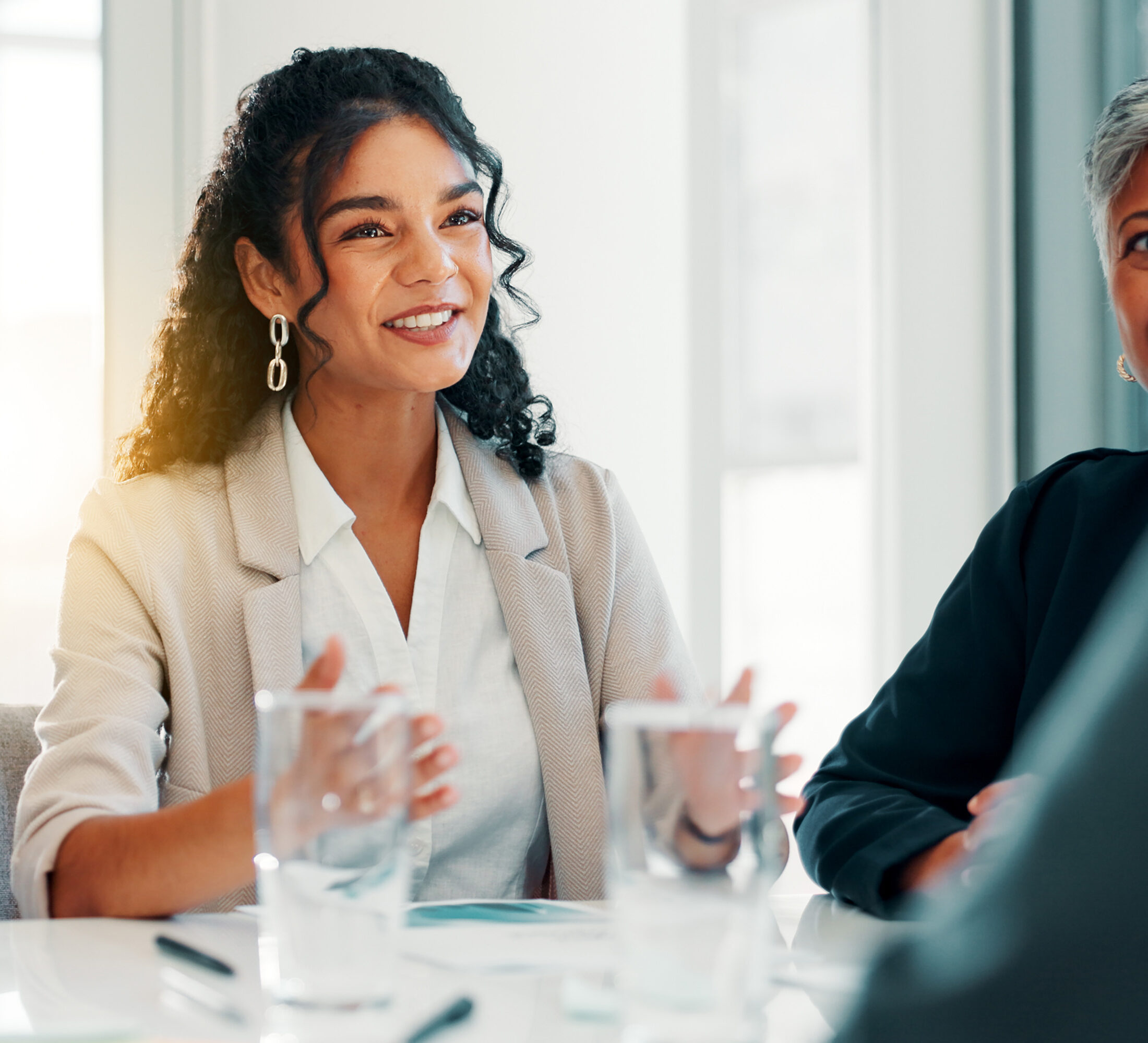 A happy female colleague wearing a white shirt, beige blazer, curly black hair which is tied back talking through Employee Benefits Advice with a client. She is using her hands to talk whilst sat down at a desk. The person she is talking to is out of shot.