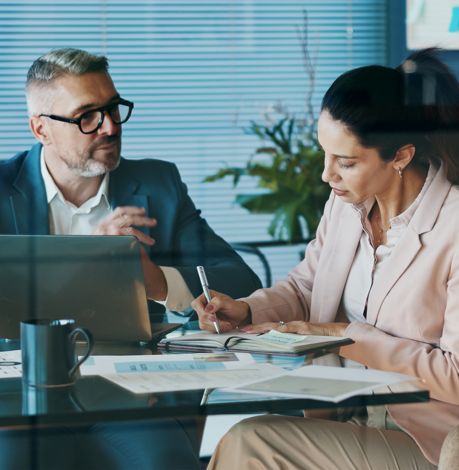 A client and employee benefits broker in a meeting discussing an employee benefits audit. The man is wearing a teal suit, talking to the client with a laptop in front of him, the client is wearing a light pink blazer and is writing down the information.