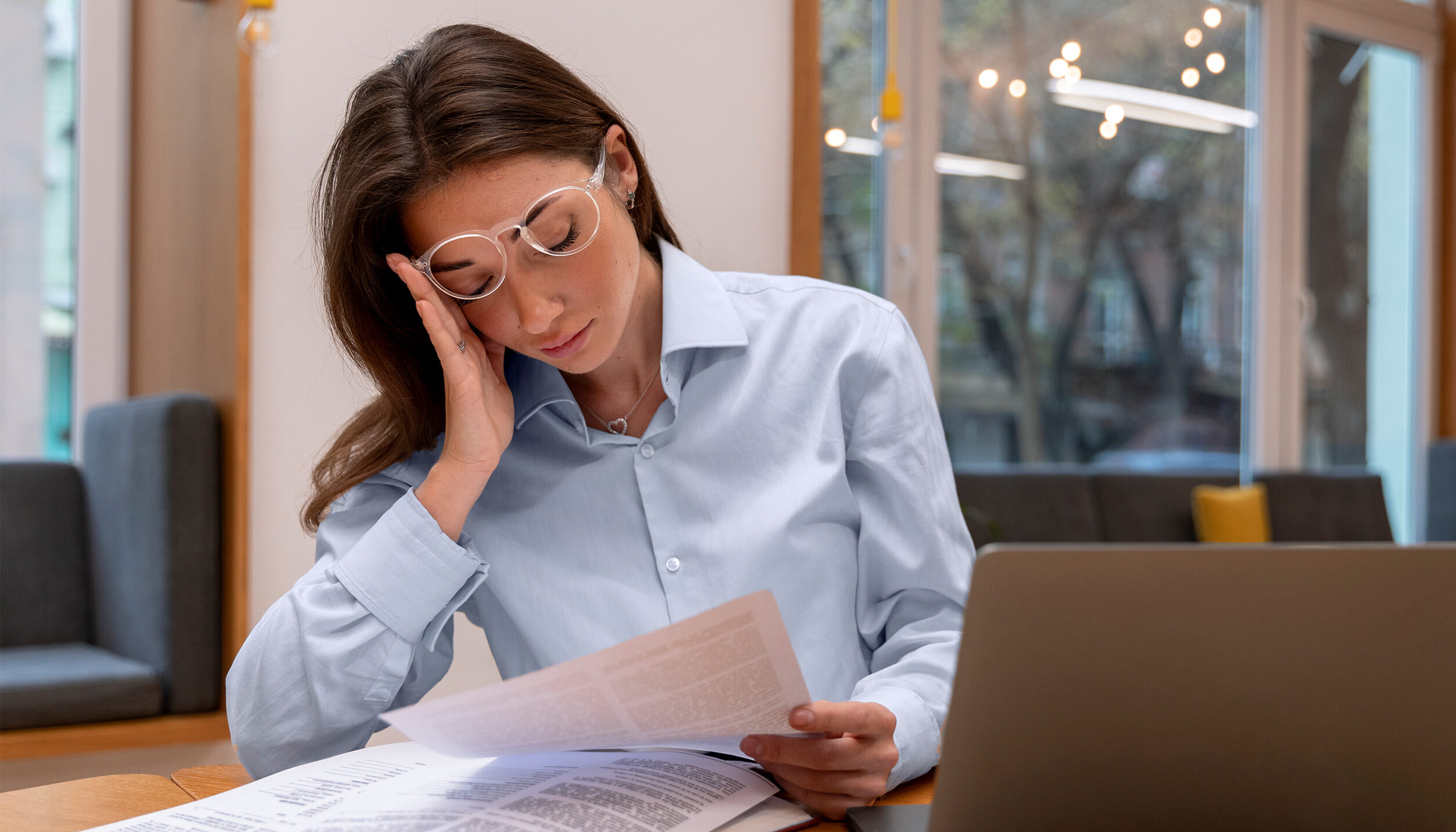 A corporate lady stressed in an office setting looking at multiple pieces of paper next to her laptop. Displaying the issue with payroll compliance