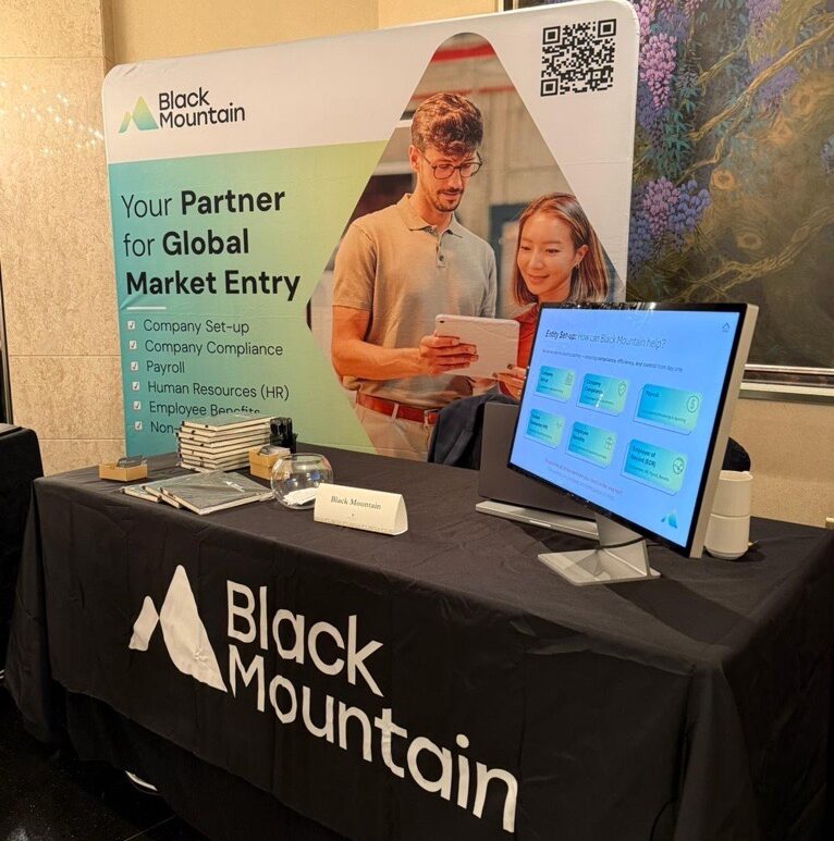 Global Market Entry: Our stall at the 2025 Singapore Business Federation Europe Conference. We have a large banner explaining how we help business gain global market entry. The banner has an image of two employees looking at an ipad together. In front of the 2m x 2m banner is a 6ft desk with black mountain branding on a table cloth, a computer with a branded slide deck presenting and a few freebies.